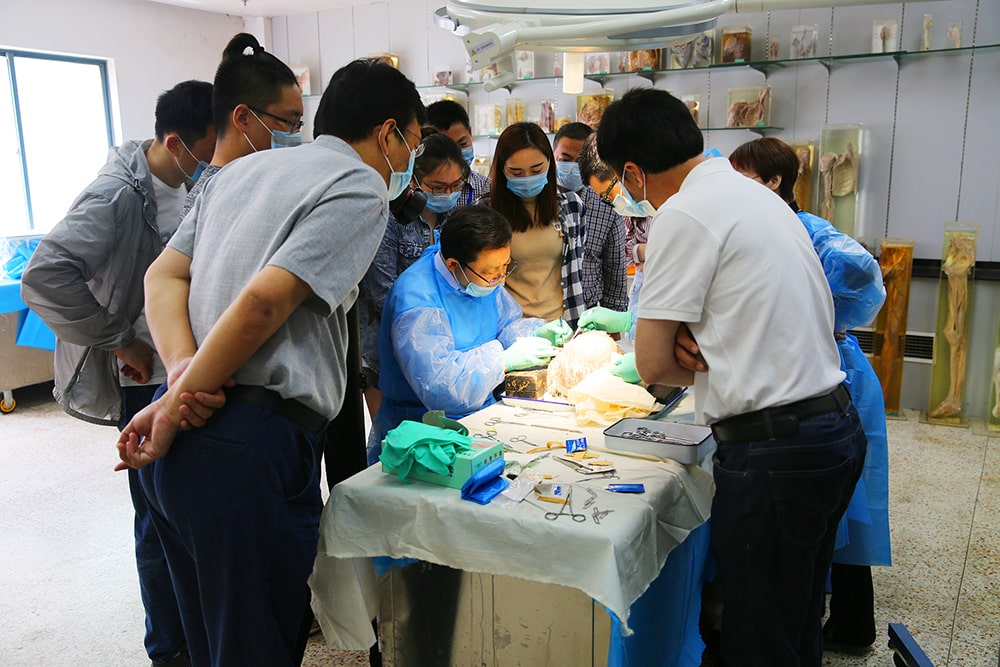A group of people, mostly in medical attire and face masks, gather closely around a central figure performing a procedure on a model at a table covered with surgical instruments.