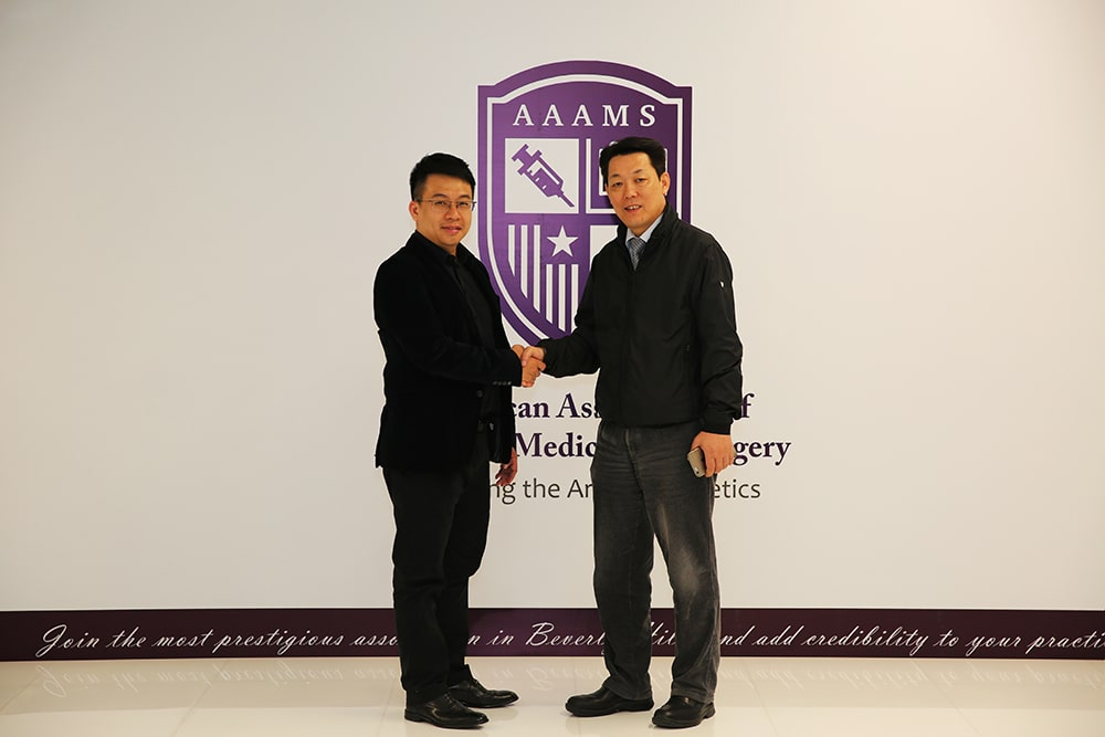 Two men shaking hands in front of a sign for the American Association of Aesthetic Medicine and Surgery (AAAMS). One wears a black suit, the other a dark jacket and casual pants.