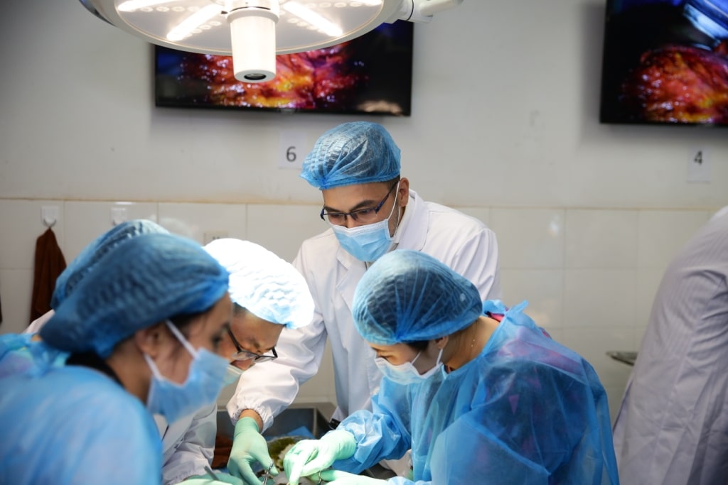 A group of medical professionals in surgical attire perform a procedure in a brightly lit operating room.