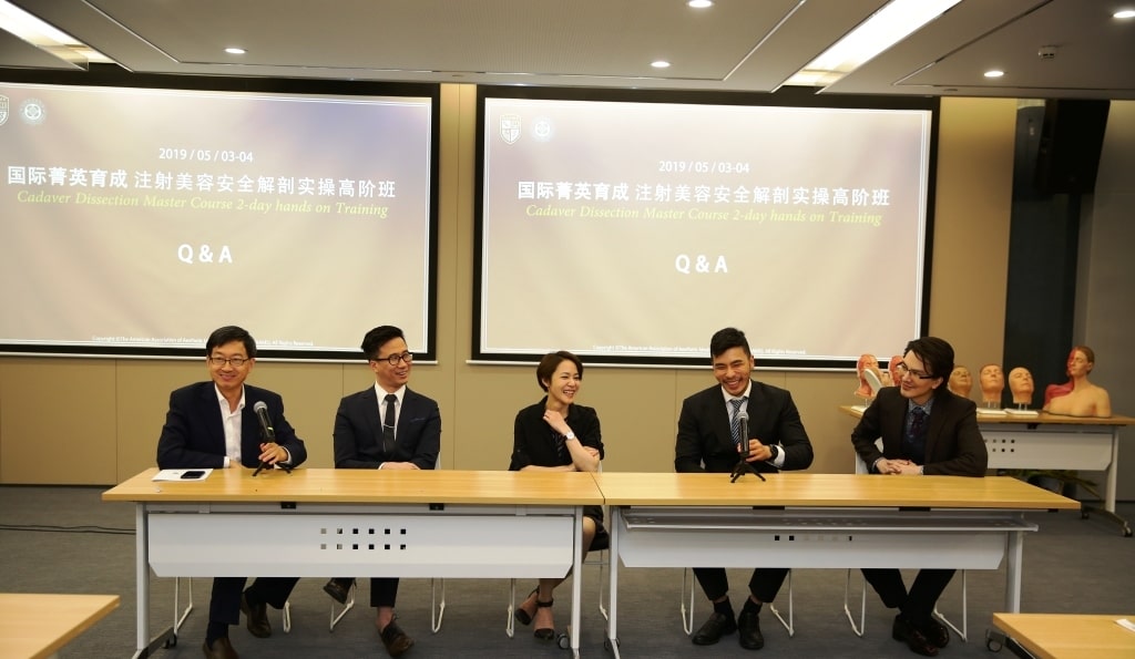 Five people are seated at a desk in front of a screen displaying the text, "Cadaver Dissection Master Course: 2-day Hands-on Training Q&A." Medical equipment and anatomical models are visible behind them.