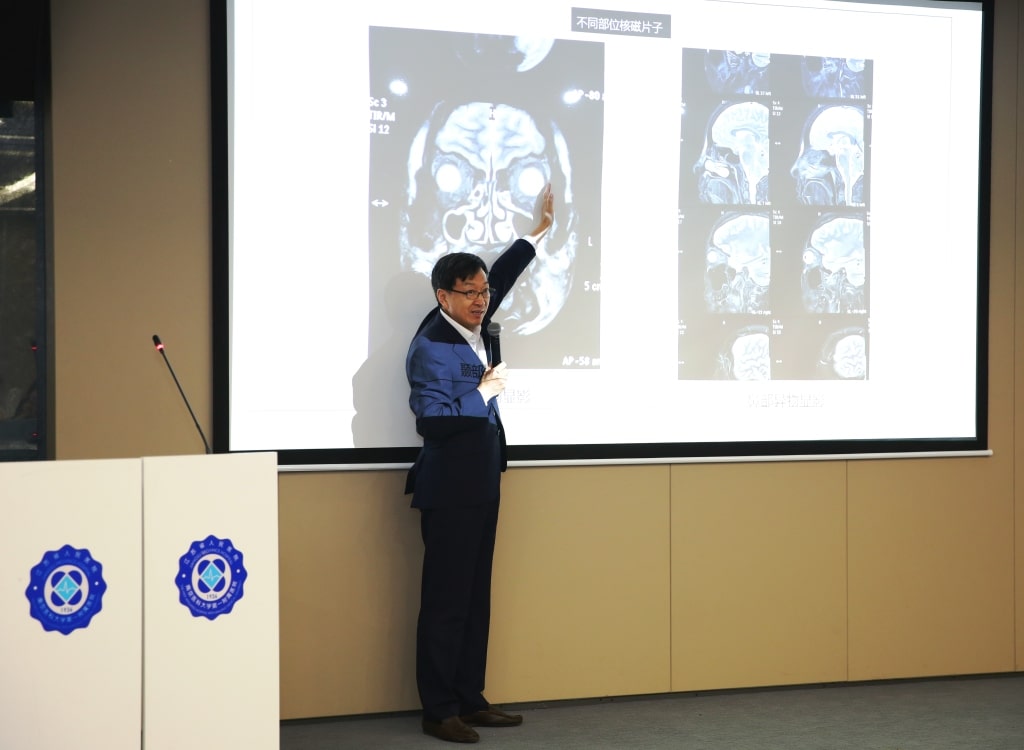 A man in a suit presents brain scan images on a large screen during a lecture, pointing to specific areas on the scans. The podium has a blue and white emblem.
