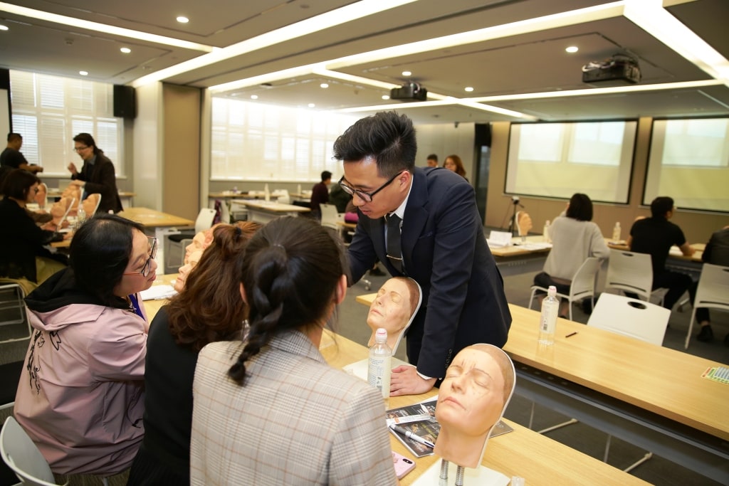 People in a classroom setting, some seated around a table with lifelike medical mannequins, engaging in a training or educational session.