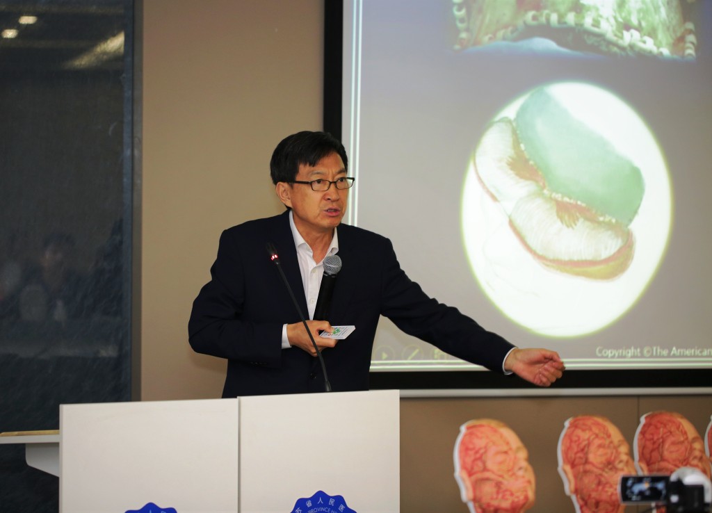 A man in a suit speaks at a podium, gesturing toward a screen displaying a diagram.