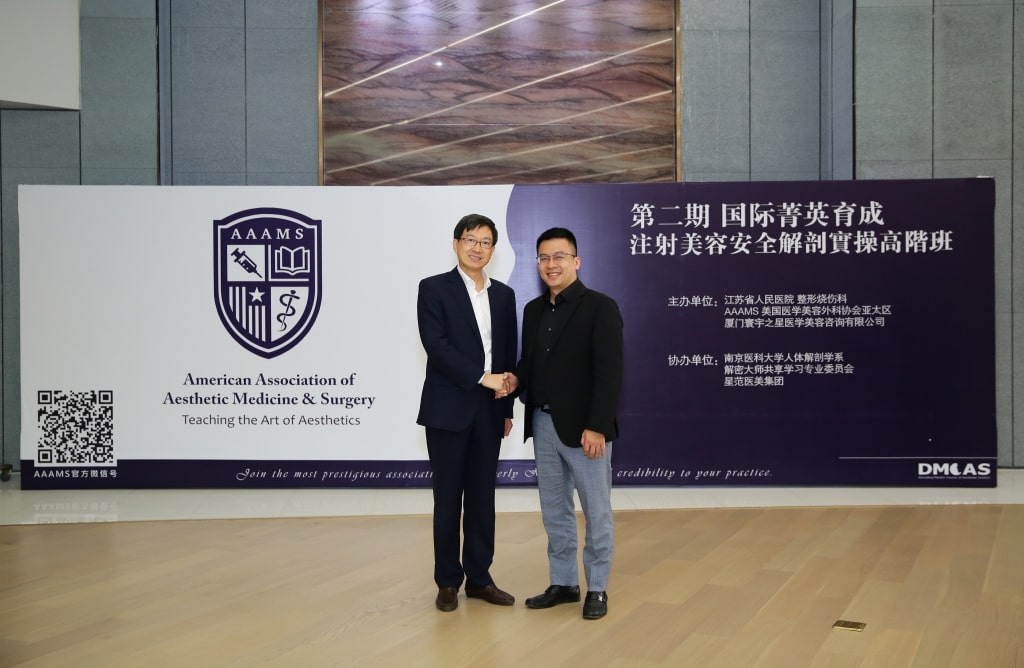 Two men stand in front of a backdrop displaying the American Association of Aesthetic Medicine & Surgery logo, shaking hands and smiling. They are dressed in business attire.