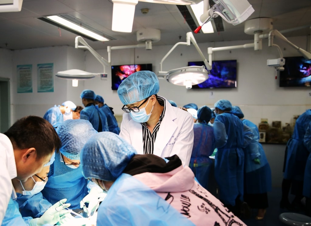 Medical professionals wearing blue surgical gowns, masks, and hair nets conduct a surgical procedure in a well-equipped operating room, illuminated by overhead lights with monitors displaying visuals.
