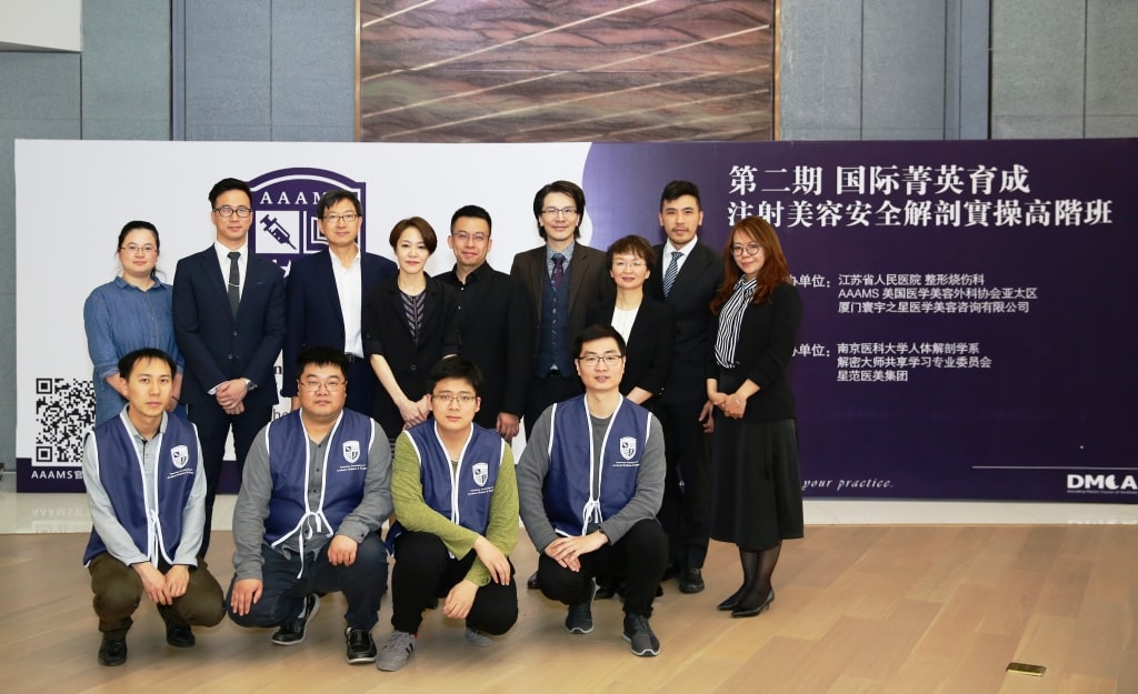 A group of 14 people, including 9 standing and 5 crouching in front, poses for a photo indoors during an event. A banner with Chinese and English text is visible in the background.