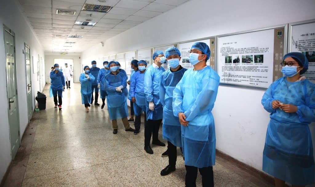 A group of healthcare workers wearing blue protective gowns, face masks, and hair covers stand lined up along a hallway with information posters on the wall.