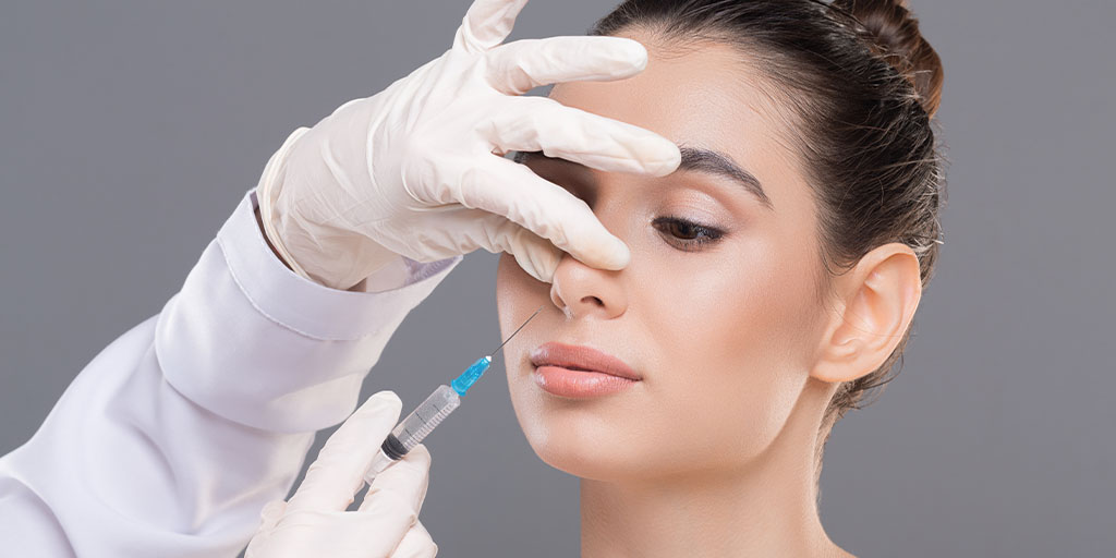 A professional wearing white gloves administers a cosmetic injection to a woman's lips, skillfully holding her nostril with one hand and guiding the syringe with the other, demonstrating expert technique that could be valuable for non-surgical rhinoplasty training.