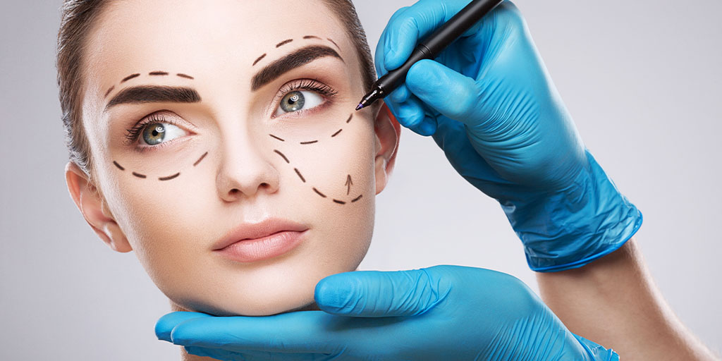 A person with surgical marker lines on their face, indicating areas for cosmetic surgery, while a gloved hand holds a marker and another hand supports their chin during an aesthetic training session for nurses.