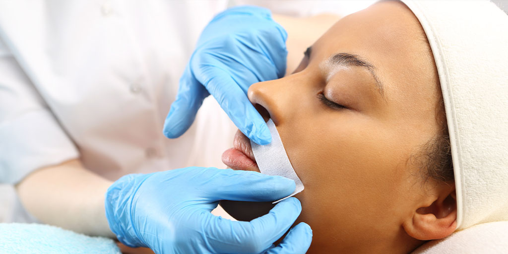 A person lies down with their eyes closed during a lip waxing procedure, as an aesthetic nurse wearing blue gloves applies a wax strip to their upper lip.