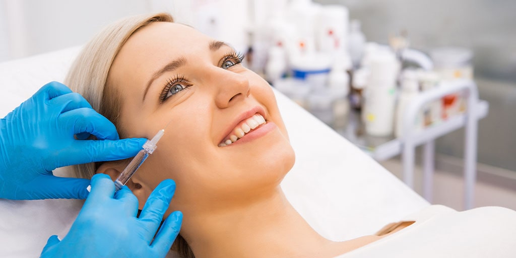 A smiling woman undergoing a facial injection procedure by a person wearing blue gloves in a medical setting, showcasing botox training in action.