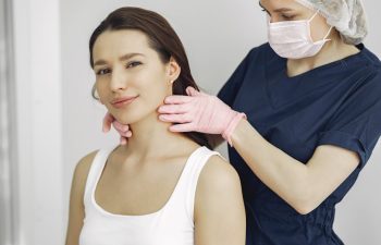 A woman wearing a white tank top receives a neck examination from a medical professional dressed in blue scrubs, a hair cap, face mask, and pink gloves.
