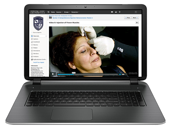 A laptop screen displays a video tutorial on facial injections, showing a woman receiving an injection in her temple from a practitioner wearing white gloves.