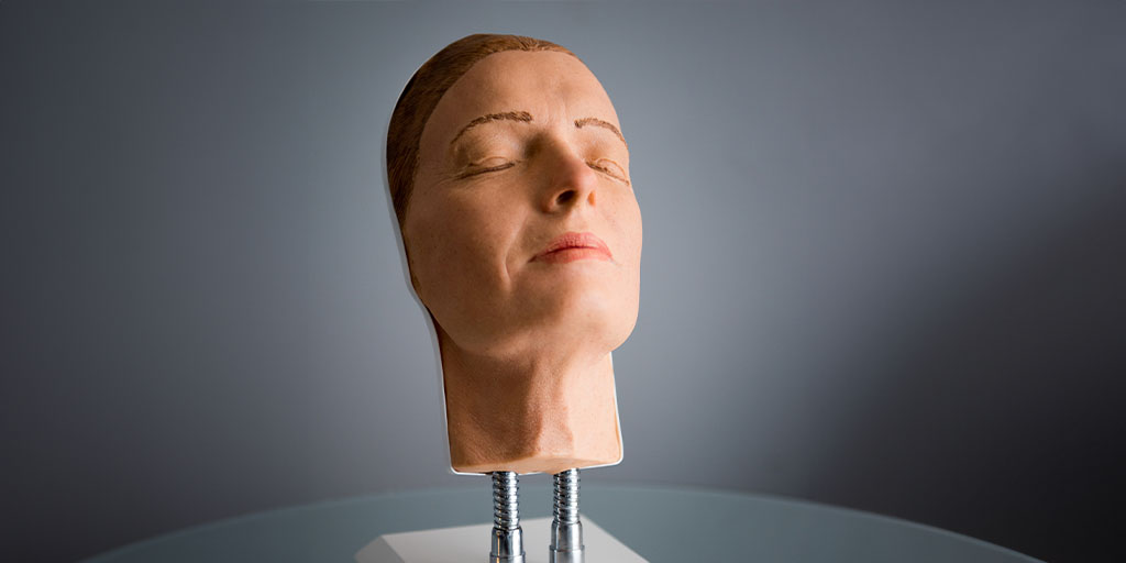 A realistic life-size head model, featuring closed eyes and natural skin tones, is mounted on a base with visible screws.