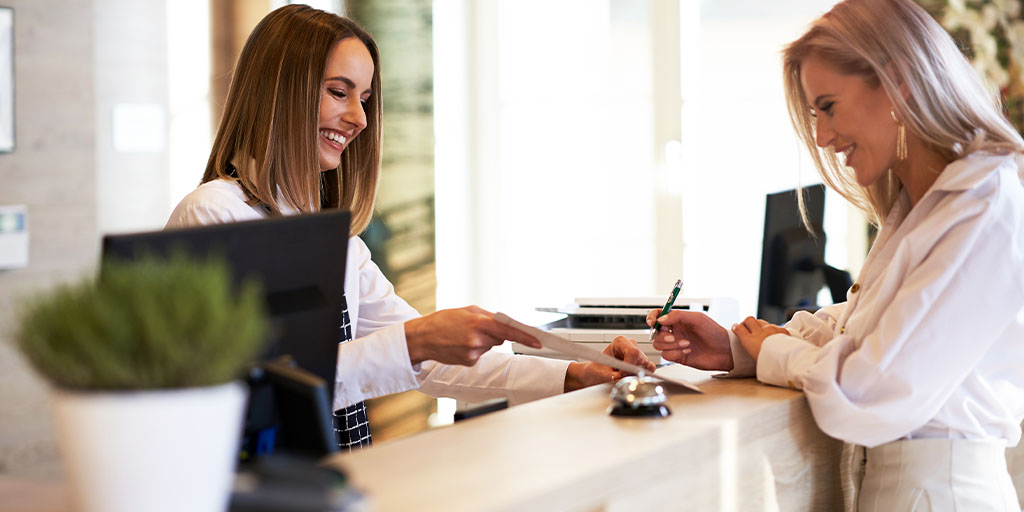 A receptionist hands a document to a smiling woman at a desk. Both women appear engaged in a pleasant interaction.