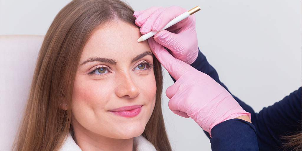 A person with long hair sits while another person wearing pink gloves marks their forehead with a white pencil.