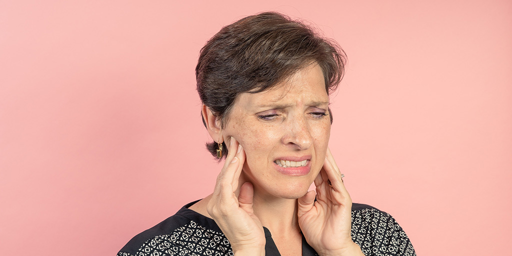 Person with short hair touching their jaw, appearing to be in pain, against a pink background.