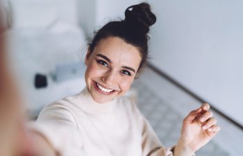 Pretty young female with big smile standing at bedroom after work with laptop and having fun taking light cheerful selfie on blurred background